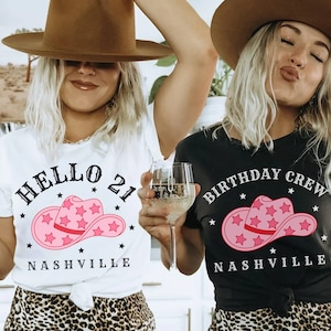 May include: Two women wearing white and black t-shirts with a pink cowboy hat graphic and the text "Hello 21 Nashville" and "Birthday Crew Nashville".