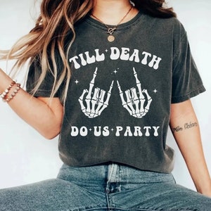 May include: A dark gray t-shirt with a white graphic print of two skeleton hands making the rock and roll hand gesture. The text "Till Death Do Us Party" is printed in white on the shirt.