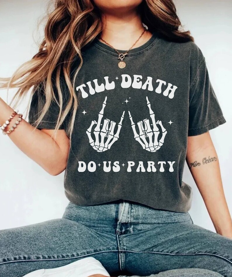 May include: A dark gray t-shirt with a white graphic print of two skeleton hands making the rock and roll hand gesture. The text "Till Death Do Us Party" is printed in white on the shirt.