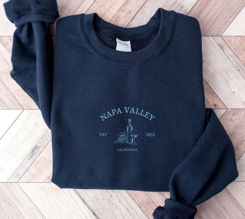 Puede incluir: Una sudadera azul marino con un dise&ntilde;o gr&aacute;fico blanco que dice "NAPA VALLEY EST 1850 CALIFORNIA" con un dibujo de una botella de vino y uvas.