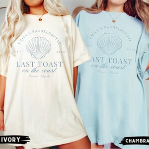K&ouml;nnte beinhalten: Zwei &uuml;bergro&szlig;e T-Shirts mit einem Muschel-Motiv und dem Text "Emily's Bachelorette Est 2024 Last Toast on the Coast Miami Florida". Die T-Shirts sind in Elfenbein und Hellblau.
