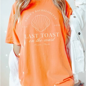 K&ouml;nnte beinhalten: Orangefarbenes T-Shirt mit wei&szlig;em Text, der "Emily's Bachelorette 2024 EST LAST TOAST on the coast Miami Florida" lautet, und einer wei&szlig;en Muschelgrafik.