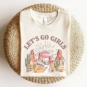 K&ouml;nnte beinhalten: Ein wei&szlig;es T-Shirt mit einem grafischen Aufdruck einer W&uuml;stenlandschaft mit Sonnenuntergang und dem Text "Let's Go Girls".