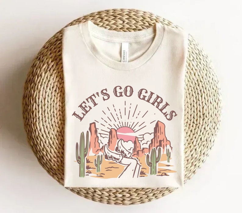 K&ouml;nnte beinhalten: Ein wei&szlig;es T-Shirt mit einem grafischen Aufdruck einer W&uuml;stenlandschaft mit Sonnenuntergang und dem Text "Let's Go Girls".