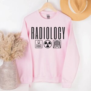 May include: A pink sweatshirt with the word "RADIOLOGY" printed in black. The word is centred on the sweatshirt and is surrounded by three black icons: a skeleton, a radiation symbol, and a chest x-ray.