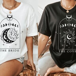 Op de afbeelding: Twee witte T-shirts met zwarte grafische ontwerpen. Het linker shirt heeft een halve maan en sterren met de tekst "THE BRIDE". Het rechter shirt heeft een zon en een maan met de tekst "THE COVEN".