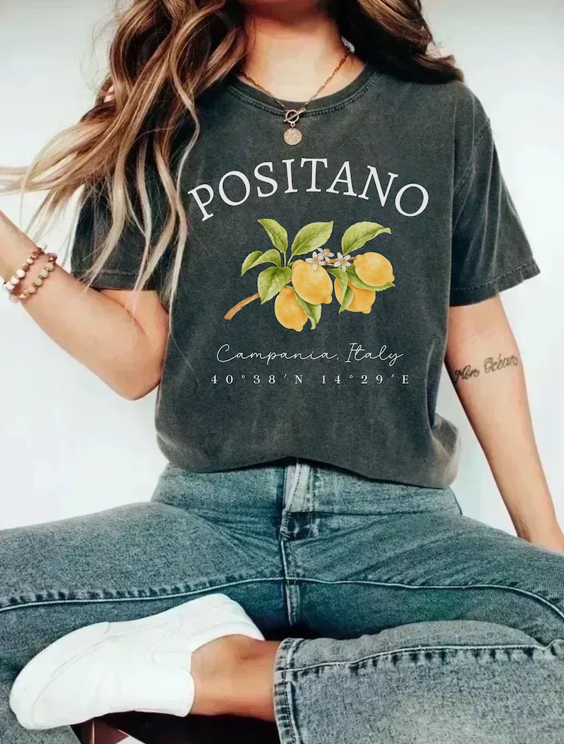 Może przedstawiać: Ciemnoszara koszulka z białym napisem "POSITANO" i ilustracją cytryn. Poniżej znajduje się napis "Campania, Italy" i wsp&oacute;łrzędne. Koszulka jest zestawiona z niebieskimi dżinsami i białymi butami.