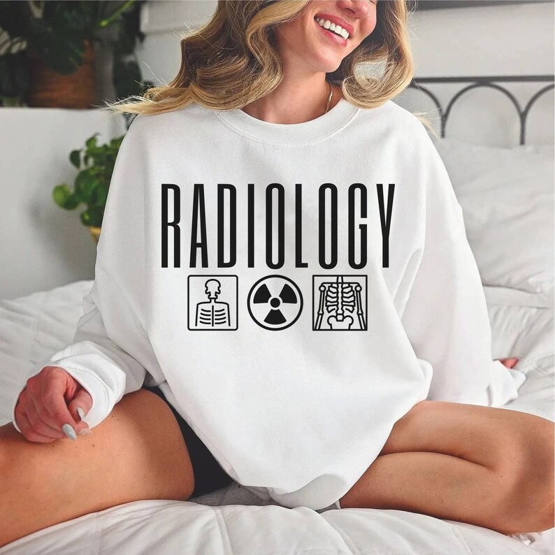 May include: White sweatshirt with the text "RADIOLOGY" in black lettering. Below the text are three black icons: a skeleton, a radiation symbol, and an x-ray image of a skeleton.