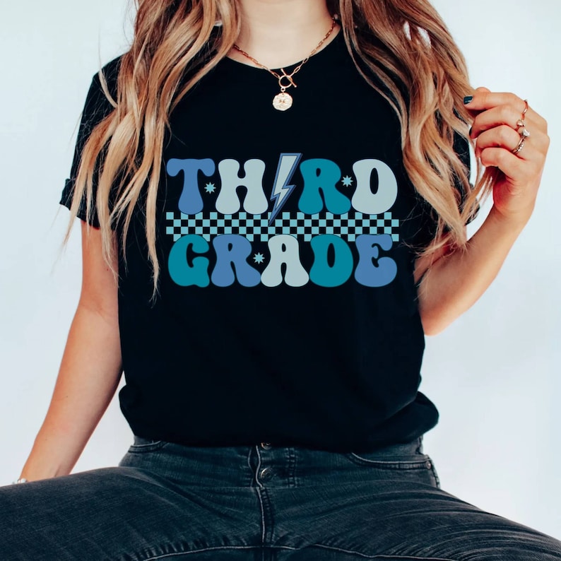 May include: A black t-shirt with a retro-style graphic that says "THIRD GRADE" in blue and teal letters with a lightning bolt between the words. The letters are outlined in white and have a checkered pattern underneath.