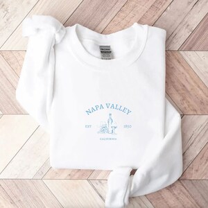 Puede incluir: Sudadera blanca con un estampado gr&aacute;fico azul que dice "Napa Valley Est. 1850 California" con un dibujo de una botella de vino y dos copas de vino.