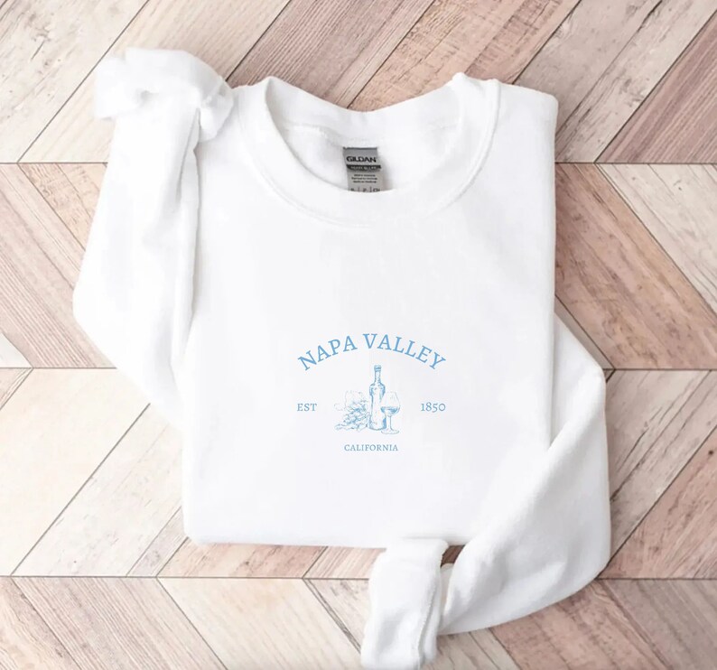 Puede incluir: Sudadera blanca con un estampado gr&aacute;fico azul que dice "Napa Valley Est. 1850 California" con un dibujo de una botella de vino y dos copas de vino.