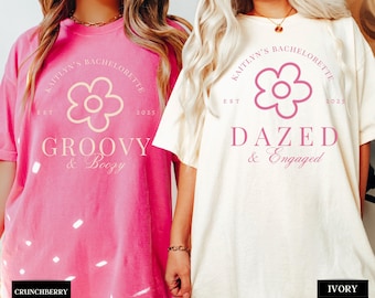 May include: Two white t-shirts with pink text and a pink flower graphic. The text on the left shirt reads "Kaitlyn's Bachelorette Est 2025 Groovy & Boozy". The text on the right shirt reads "Kaitlyn's Bachelorette Est 2025 Dazed & Engaged".