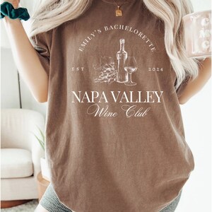 May include: A brown t-shirt with white text that reads "Emily's Bachelorette Est 2024 Napa Valley Wine Club". The shirt features a graphic of a bottle of wine, a glass of wine, and grapes.