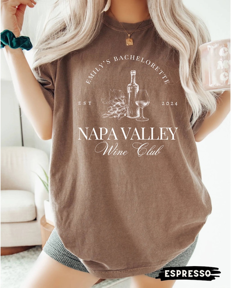 May include: A brown t-shirt with white text that reads "Emily's Bachelorette Est 2024 Napa Valley Wine Club". The shirt features a graphic of a bottle of wine, a glass of wine, and grapes.