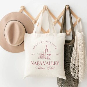 May include: A white tote bag with a red design that says "Emily's Bachelorette Est 2024 Napa Valley Wine Club". The design also includes a bottle of wine, a glass of wine, and grapes.