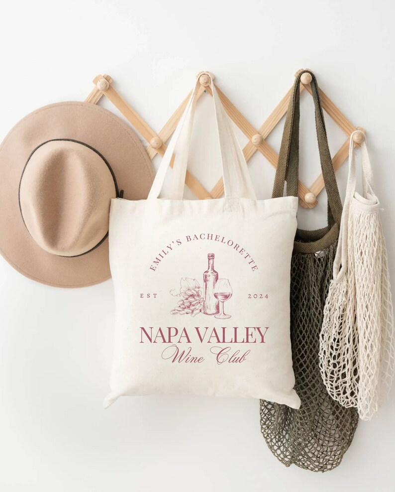May include: A white tote bag with a red design that says "Emily's Bachelorette Est 2024 Napa Valley Wine Club". The design also includes a bottle of wine, a glass of wine, and grapes.