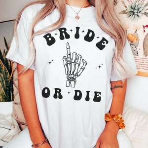 May include: White t-shirt with a black graphic of a skeleton hand with a ring on its finger. The text "BRIDE OR DIE" is written in black letters with a retro font.