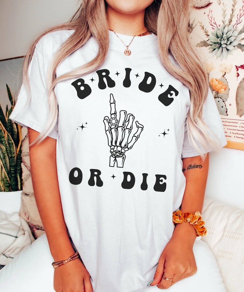May include: White t-shirt with a black graphic of a skeleton hand with a ring on its finger. The text "BRIDE OR DIE" is written in black letters with a retro font.