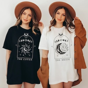 Op de afbeelding: Twee vrouwen dragen zwarte en witte T-shirts met maan- en stermotieven. Het zwarte T-shirt zegt "The Coven" en het witte T-shirt zegt "The Bride".