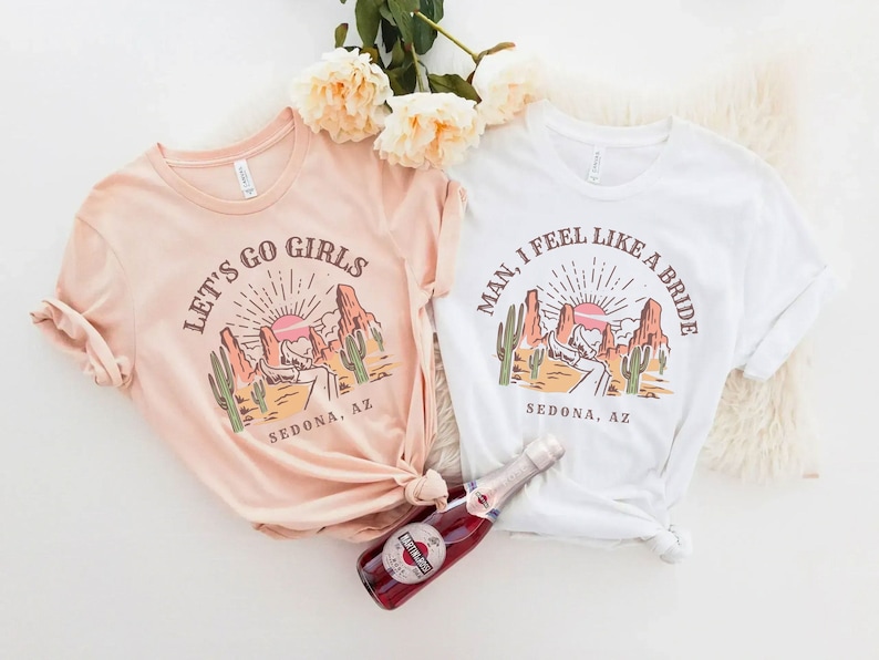 May include: Two t-shirts with a desert landscape design. One shirt is peach colored and says "Let's Go Girls Sedona, AZ". The other shirt is white and says "Man, I Feel Like A Bride Sedona, AZ".