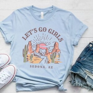 May include: Light blue t-shirt with a graphic of a desert landscape with a sunset and the text "Let's Go Girls Sedona, AZ".