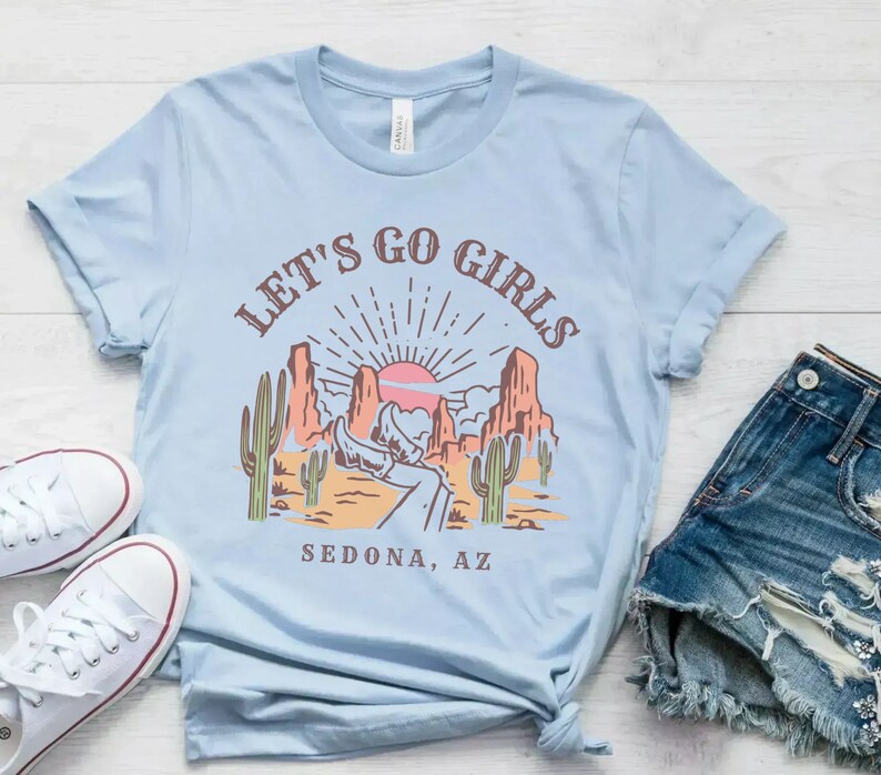 May include: Light blue t-shirt with a graphic of a desert landscape with a sunset and the text "Let's Go Girls Sedona, AZ".