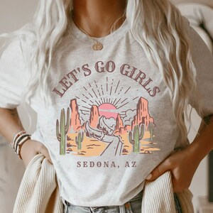 May include: A white t-shirt with a graphic print of a desert landscape with a sunset and the text "Let's Go Girls Sedona, AZ".