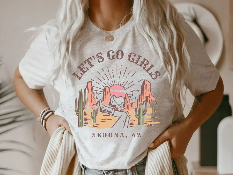 May include: A white t-shirt with a graphic print of a desert landscape with a sunset and the text "Let's Go Girls Sedona, AZ".