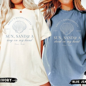 May include: Two white t-shirts with blue text and a seashell graphic. The text on the left shirt reads "Emily's Bachelorette Est 2024 Sun, Sand & A ring on my hand Miami Florida". The text on the right shirt reads "Emily's Bachelorette Est 2024 Sun, Sand & A drink in my hand Miami Florida". The left shirt is ivory and the right shirt is blue jean.