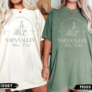 May include: Two oversized t-shirts with a vintage-inspired design. The shirts are in ivory and moss green colors. The design features a bottle of wine, a glass of wine, and grapes with the text "Emily's Bachelorette Est 2024 Napa Valley Wine Club".