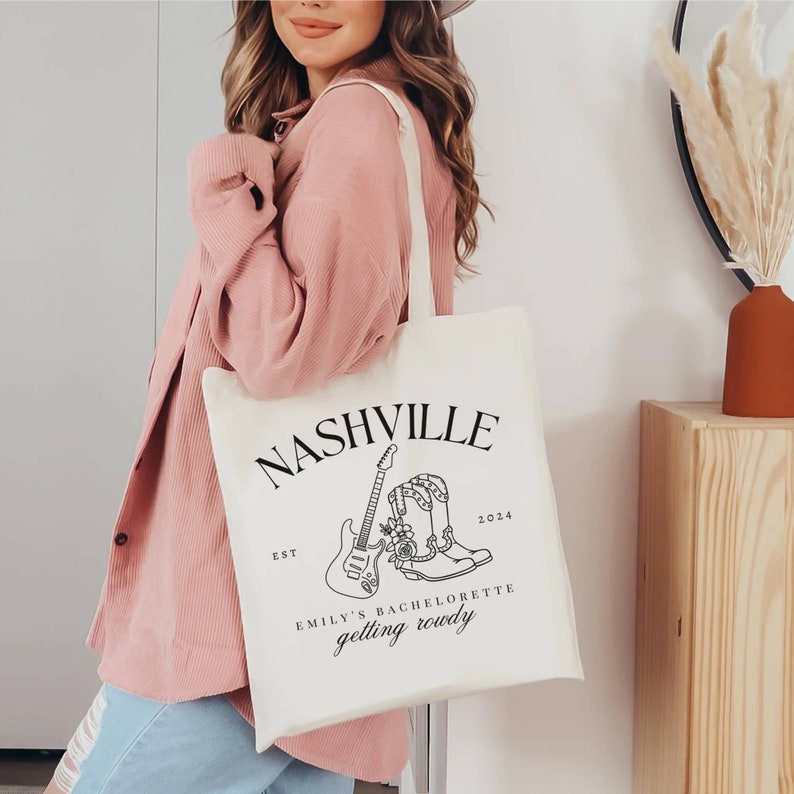 May include: White canvas tote bag with black text and graphics. The text reads "Nashville Est 2024 Emily's Bachelorette getting rowdy". The graphics are a guitar and a pair of cowboy boots.