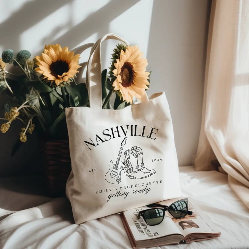 May include: A white canvas tote bag with black text that reads "Nashville Est 2024 Emily's Bachelorette getting rowdy". The bag features a black and white illustration of a guitar and cowboy boots with flowers.