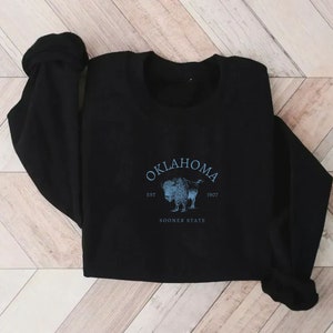 Puede incluir: Sudadera negra con un estampado gr&aacute;fico azul y blanco de un b&uacute;falo con el texto "Oklahoma Est 1907 Sooner State"