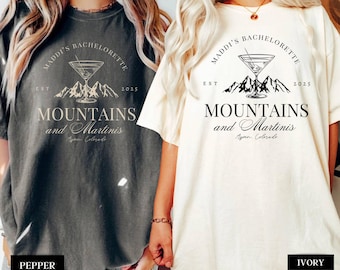 May include: Two t-shirts with a mountain and martini graphic. The text on the shirts reads "Maddi's Bachelorette Est 2025 Mountains and Martinis Aspen Colorado". The shirts are available in pepper and ivory.