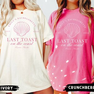 K&ouml;nnte beinhalten: Zwei &uuml;bergro&szlig;e T-Shirts mit einem rosa Muschel-Motiv und dem Text "Emily's Bachelorette Est 2024 Last Toast on the Coast Miami, Florida" auf der Vorderseite. Die T-Shirts sind in den Farben Elfenbein und Crunchberry.