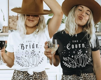 May include: Two women wearing white and black t-shirts with the text "The Bride" and "The Coven" in a gothic font. Both shirts feature a design of crystals and leaves. The women are wearing brown cowboy hats and holding wine glasses.