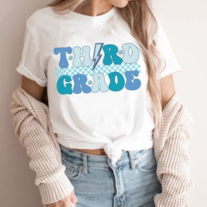 May include: A white t-shirt with a blue and white checkered pattern and the text "THIRD GRADE" in a retro font with a lightning bolt and stars.