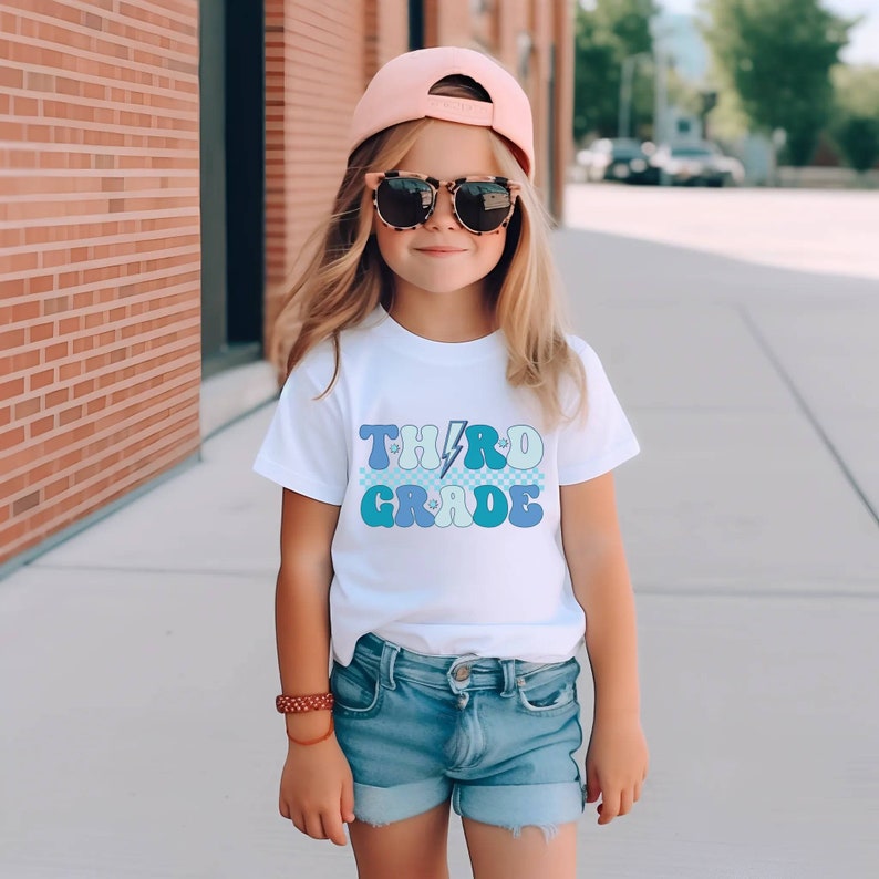 May include: A white t-shirt with a blue and light blue graphic that says "THIRD GRADE" with a lightning bolt in the middle. The shirt is being worn by a child wearing a pink baseball cap and sunglasses.