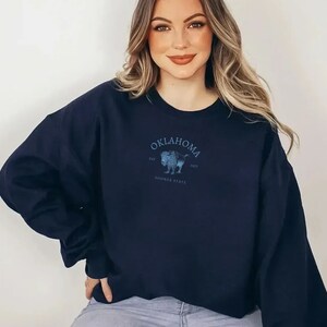 Puede incluir: Una sudadera azul marino con un gr&aacute;fico de un b&uacute;falo y el texto "Oklahoma Est. 1907 Sooner State".