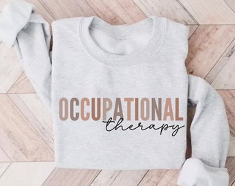 May include: A light gray sweatshirt with the words "OCCUPATIONAL therapy" printed across the front in a gradient of brown and black script. The sleeves are rolled up, revealing a white interior.
