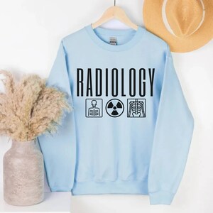 May include: Light blue sweatshirt with the word "RADIOLOGY" printed in black. The sweatshirt has three black icons below the word: a skeleton, a radiation symbol, and an x-ray image.