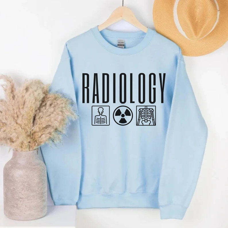 May include: Light blue sweatshirt with the word "RADIOLOGY" printed in black. The sweatshirt has three black icons below the word: a skeleton, a radiation symbol, and an x-ray image.