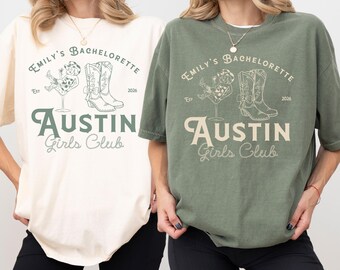 May include: Two t-shirts, one cream and one olive green, feature the text "Emily's Bachelorette Austin Girls Club" with a graphic of a cocktail, cowboy boots, and the year 2026. The shirts are casual and have short sleeves.