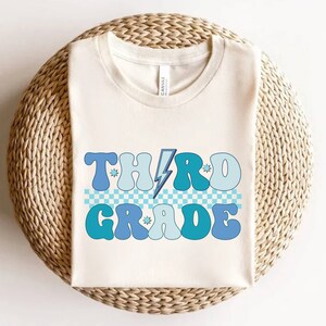 May include: A white t-shirt with a blue and light blue graphic that says "THIRD GRADE" in a retro font with a lightning bolt and stars.