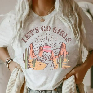 K&ouml;nnte beinhalten: Ein wei&szlig;es T-Shirt mit einem grafischen Aufdruck einer W&uuml;stenlandschaft mit Sonnenuntergang und dem Text "Let's Go Girls".