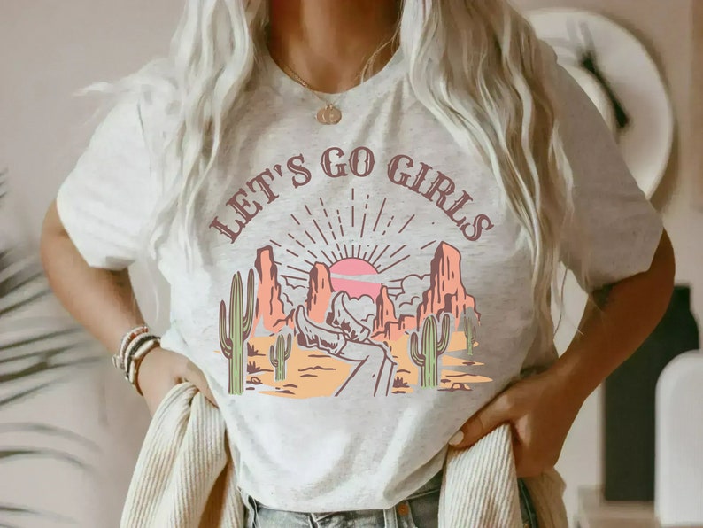 K&ouml;nnte beinhalten: Ein wei&szlig;es T-Shirt mit einem grafischen Aufdruck einer W&uuml;stenlandschaft mit Sonnenuntergang und dem Text "Let's Go Girls".