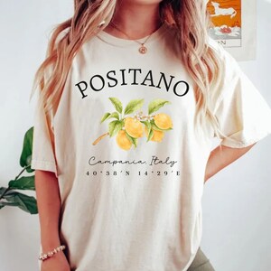 Może przedstawiać: Kremowa koszulka z czarnym napisem "POSITANO", nad akwarelową ilustracją cytryn na gałęzi. Poniżej napis "Campania, Italy" i wsp&oacute;łrzędne 40&deg;38'N 14&deg;29'E.