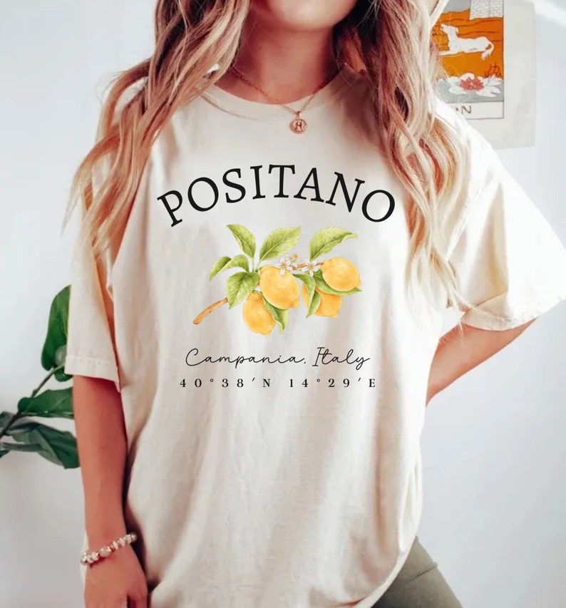 Może przedstawiać: Kremowa koszulka z czarnym napisem "POSITANO", nad akwarelową ilustracją cytryn na gałęzi. Poniżej napis "Campania, Italy" i wsp&oacute;łrzędne 40&deg;38'N 14&deg;29'E.