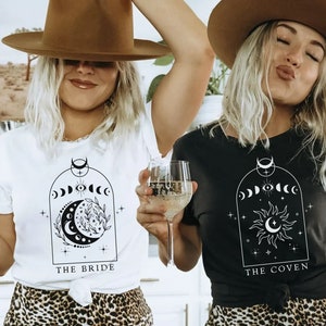 Op de afbeelding: Twee mensen dragen zwarte en witte T-shirts met een maan- en zonontwerp. De shirts zeggen "The Bride" en "The Coven".