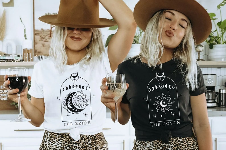 Op de afbeelding: Twee mensen dragen zwarte en witte T-shirts met een maan- en zonontwerp. De shirts zeggen "The Bride" en "The Coven".